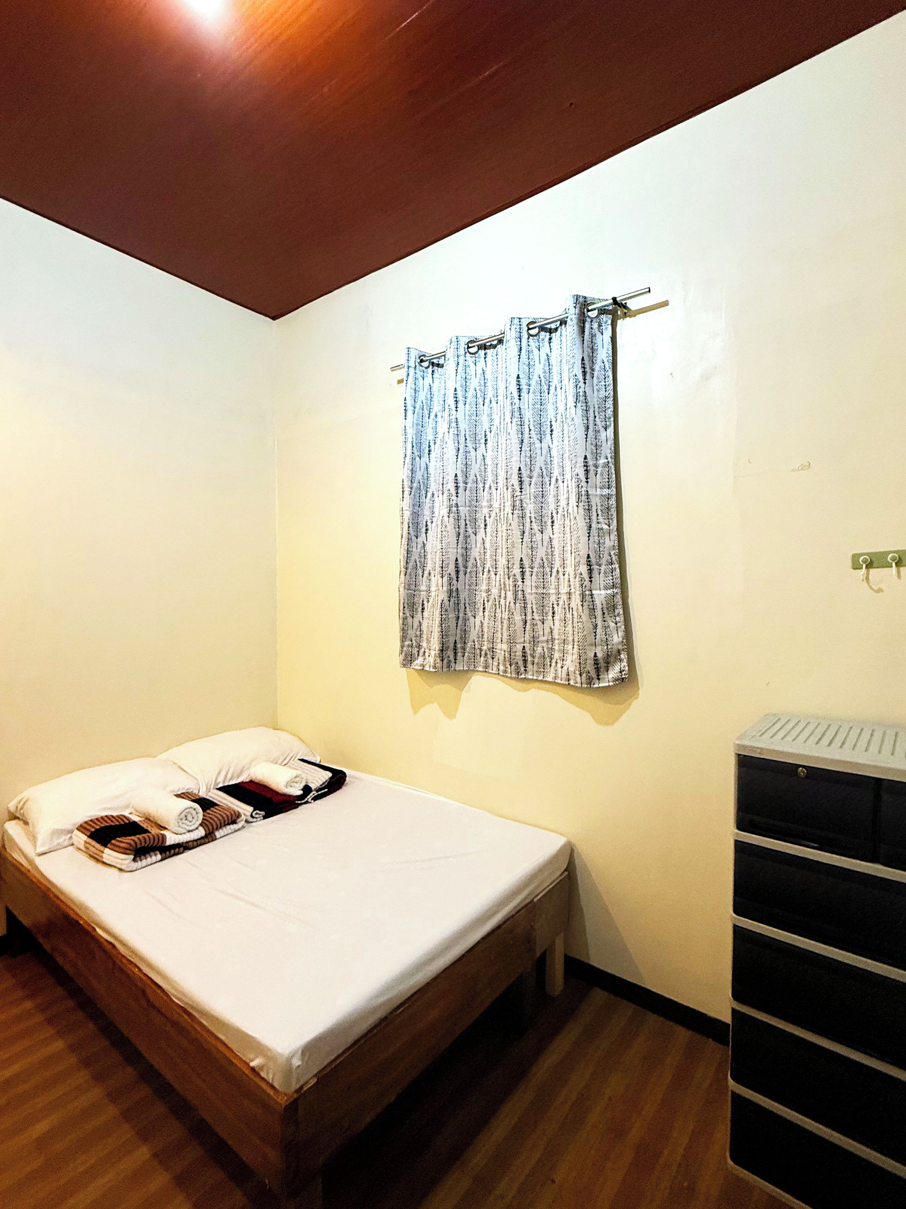 A single bed with pillows and a blanket, a wooden headboard, a wall-mounted curtain, and a black dresser with drawers.
