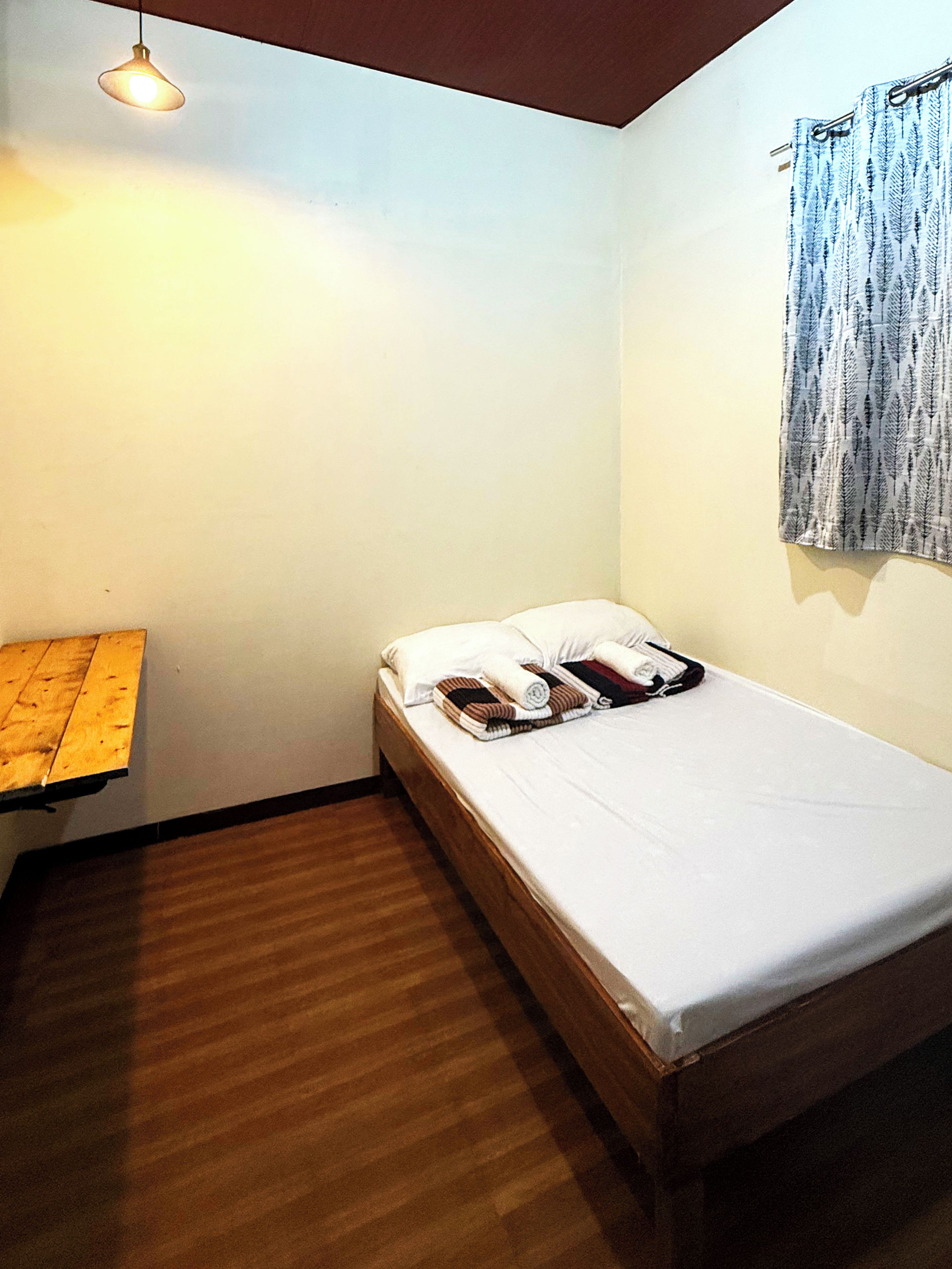 A small, single-bed room with a wooden floor, a bedside table, a hanging light fixture, and a window with a curtain.