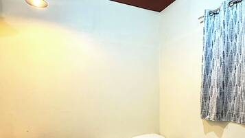 A small, single-bed room with a wooden floor, a bedside table, a hanging light fixture, and a window with a curtain.