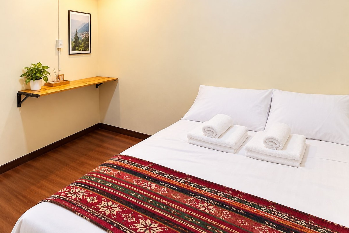 Quarto de hotel com cama, almofadas brancas, toalha branca enrolada, manta vermelha estampada, prateleira pequena com uma planta e quadro pendurado na parede.