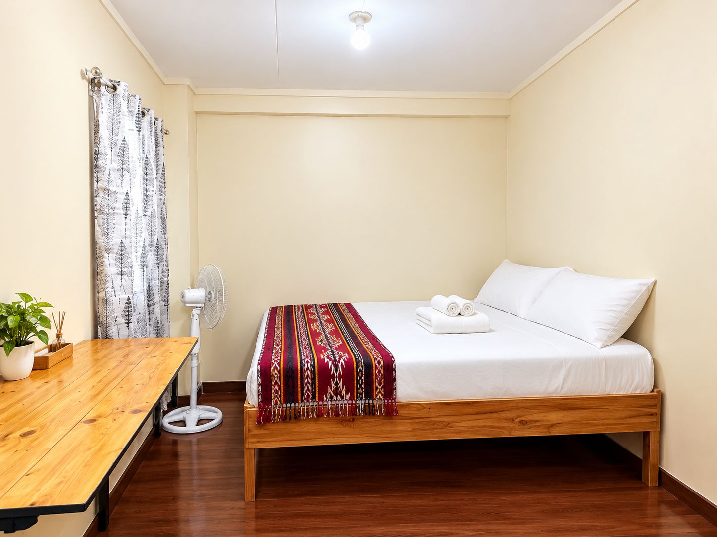 A single-bed room with a wooden floor, a bedside table with a plant, a white fan, and a red patterned blanket.