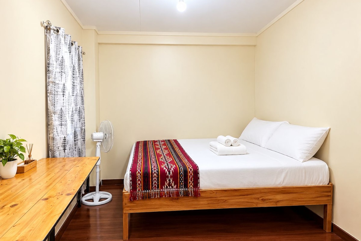 Quarto individual com uma cama, piso de madeira, mesa de cabeceira com uma planta, ventoinha branca e manta vermelha estampada.