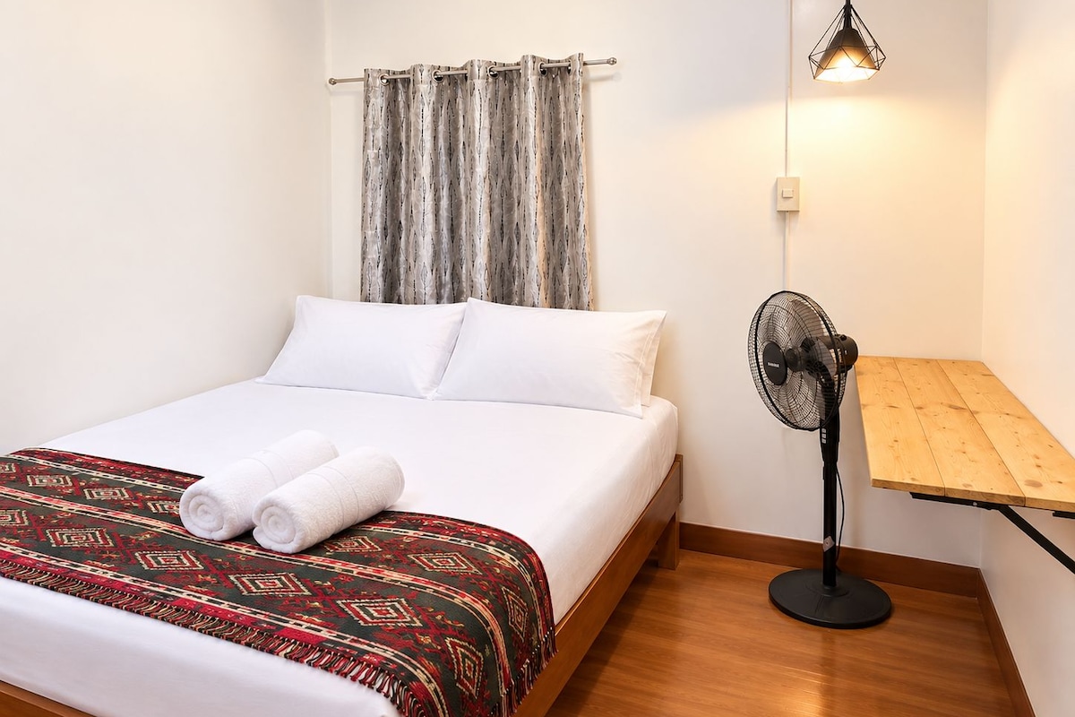 Cama bem arrumada com lençóis brancos, manta estampada e toalhas dobradas. Um ventilador de chão está ao lado da cama, e uma pequena mesa de madeira encosta na parede.