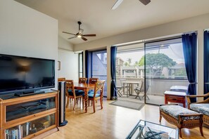 Ein Wohnzimmer mit Flachbildfernseher, Holzmöbeln, einem Glastisch und einem Balkon mit Blick auf Gebäude und Bäume.