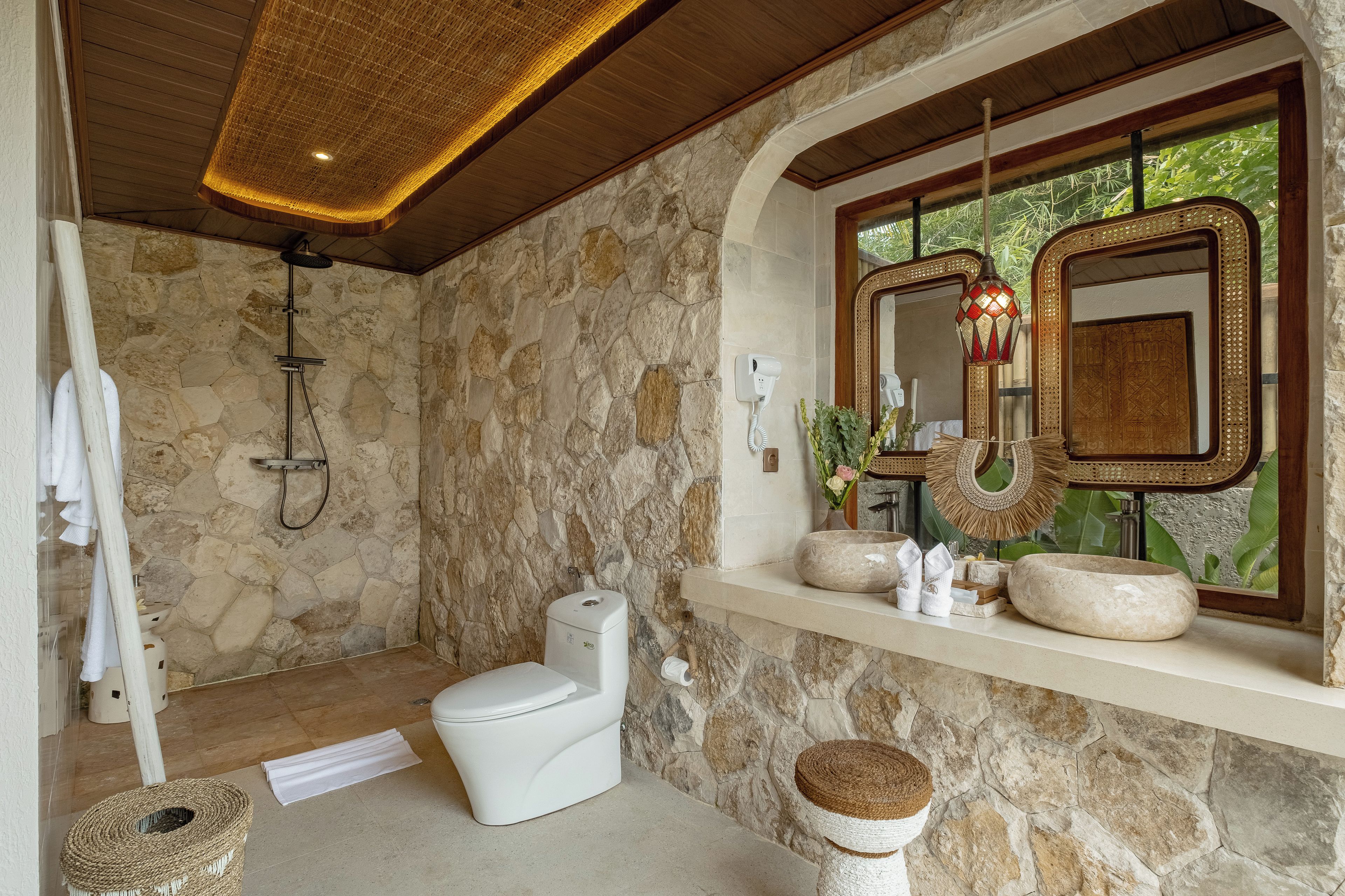 A bathroom with a stone wall, a shower, a toilet, a sink with a mirror, and a window with a view of greenery.