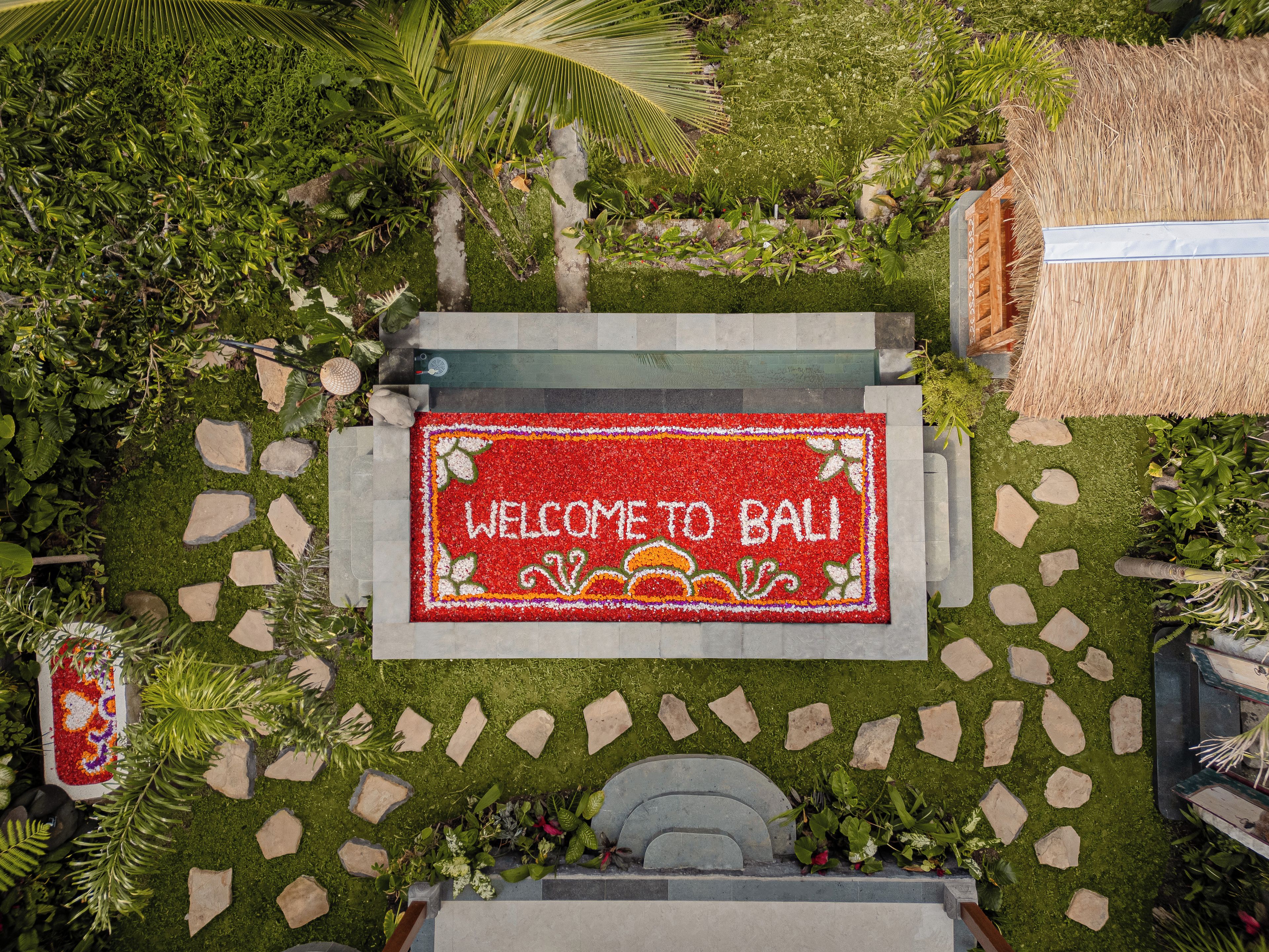 Tapete de boas-vindas com a inscrição 'WELCOME TO BALI', cercado por um jardim com diversas plantas e um caminho de pedra.