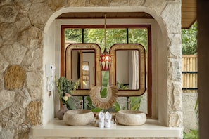 A bathroom with a stone wall, a wooden door, a mirror, a hanging light fixture, and two stone basins. - Dedari Sebatu Villa (Gianyar)