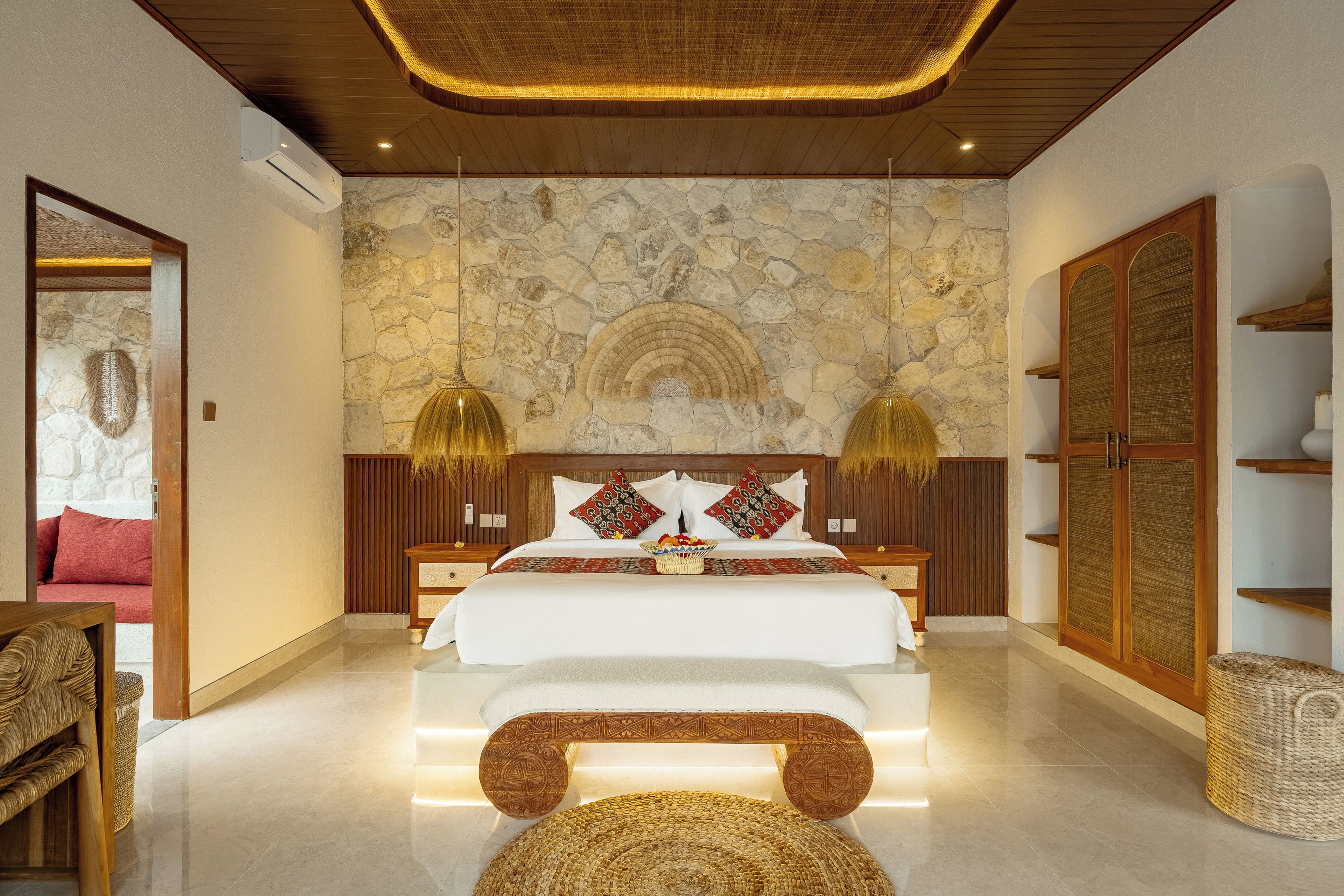 Quarto com parede de pedra, cama com almofadas estampadas, guarda-roupa de madeira e um banco de vime.
