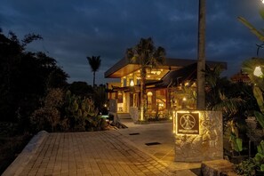 Un resort bien iluminado con fachada de piedra, área de piscina y exuberante vegetación tropical.