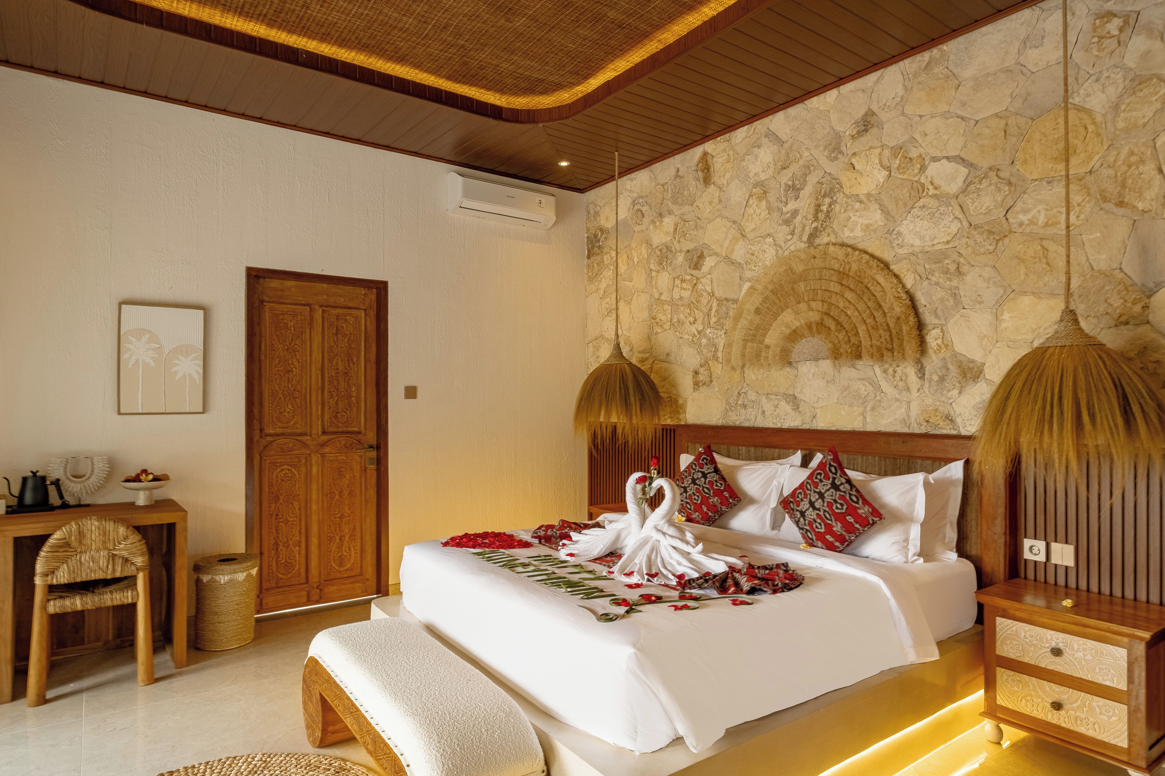 A well-appointed bedroom with a stone wall, a wooden door, a bedside table, a bench, and a bed with decorative swans and flowers.