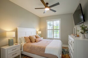 Ein Schlafzimmer mit Bett, Nachttischen, Deckenventilator, Fernseher und einem Fenster mit Jalousien.