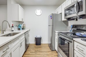 A modern kitchen with white cabinetry, stainless steel appliances, a clock on the wall, and a trash bin. - Gateway to UF & Dtwn Gainesville! Condo w/ Pool (Gainesville)