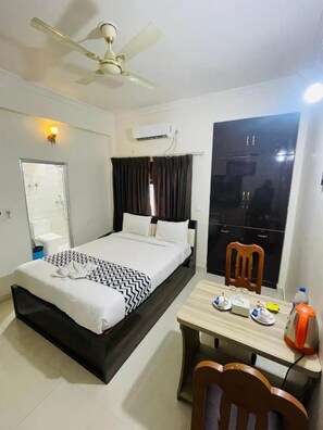 A hotel room with a bed, a desk, a chair, a ceiling fan, an air conditioning unit, a mirror, and a window with curtains. - Hotel Janakpur Inn (Janakpur)
