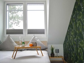 A bedroom with a bed, a bedside table with a lamp, a window with blinds, and a wall with a tropical leaf pattern. - Dream Crown Sky Blue (Schortens)