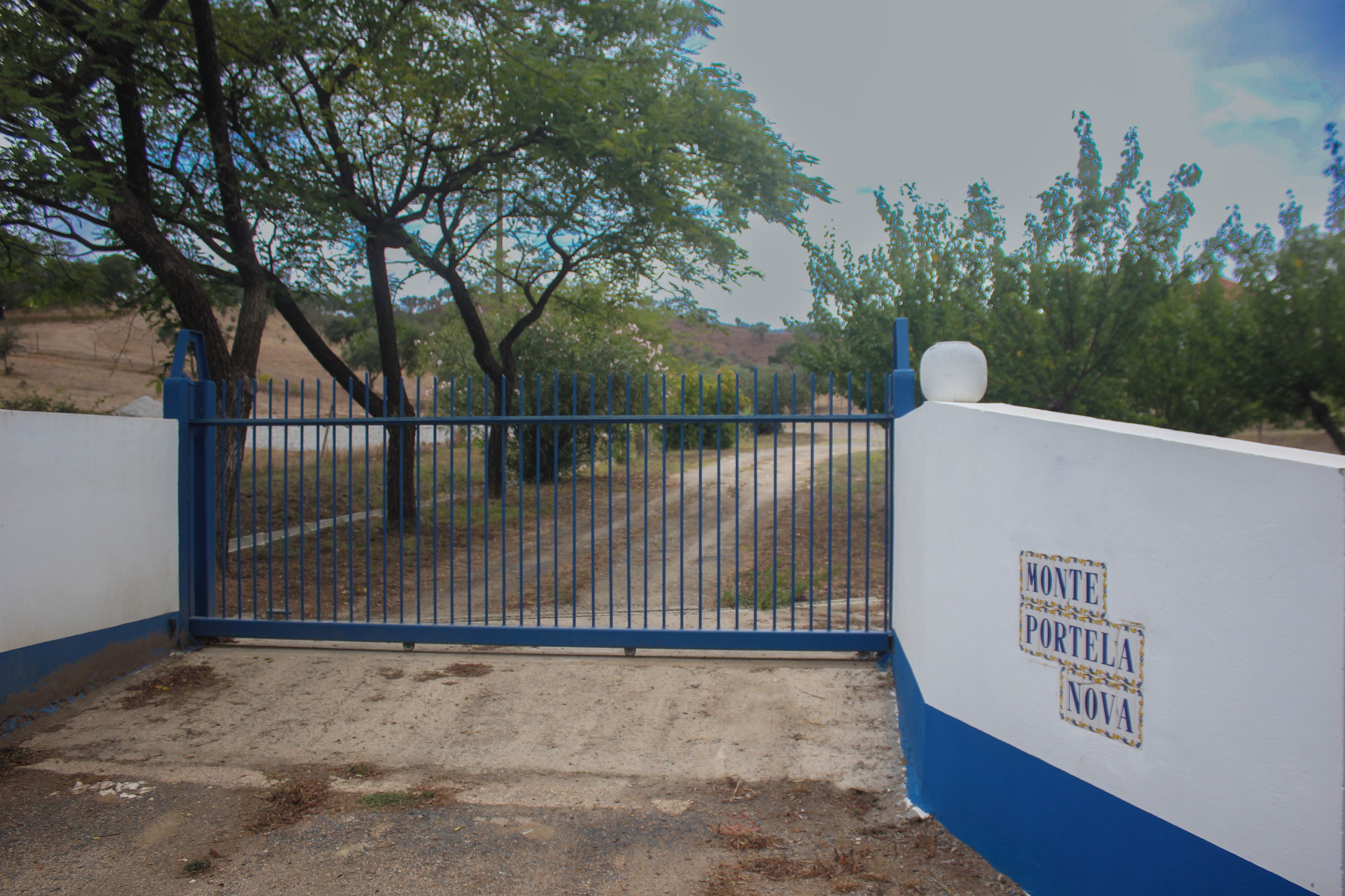 A gated entrance with a blue metal gate, a white wall with a blue stripe, and a sign that reads 'MONTE PORTELA NOVA'.