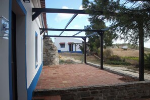 A covered patio area with a stone wall, a view of a building with a red-tiled roof, and a clear sky.