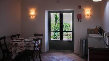 Un coin repas avec une table dressée pour quatre personnes, une porte donnant sur un espace verdoyant et un coin cuisine équipé d’une poubelle et d’un grille-pain.