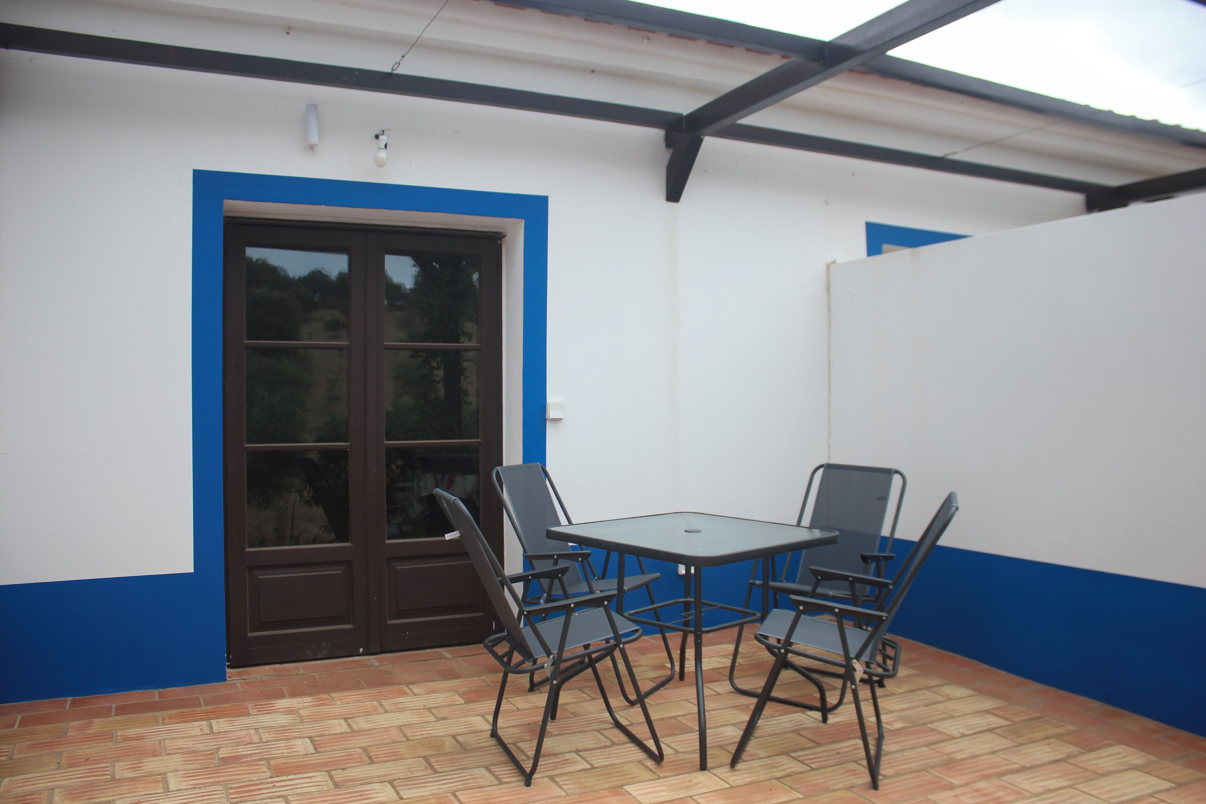 Un espace extérieur aménagé en terrasse, avec une table et quatre chaises, une porte et un sol carrelé.