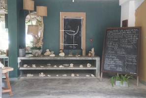 A room with a chalkboard displaying services and prices, a shelf with pottery, and a window with a view.