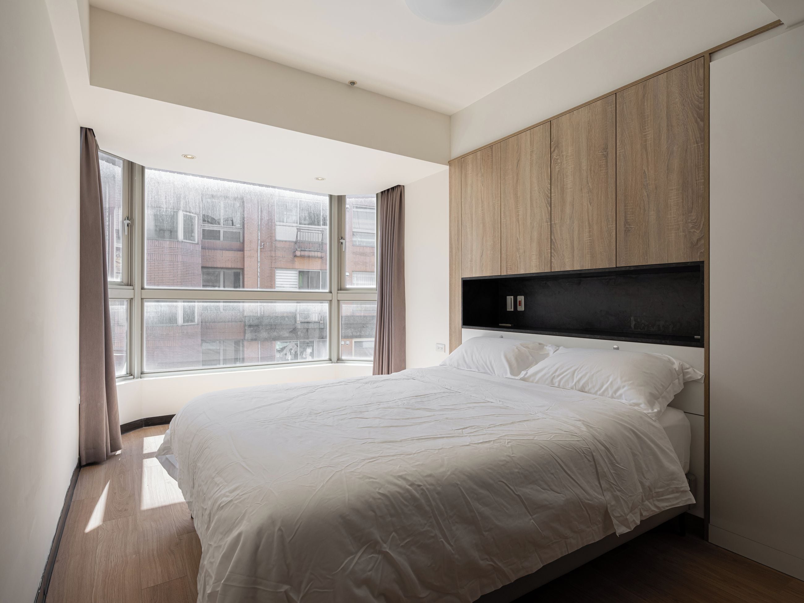 A modern bedroom with a large window, a bed with white bedding, and wooden paneling on the walls.