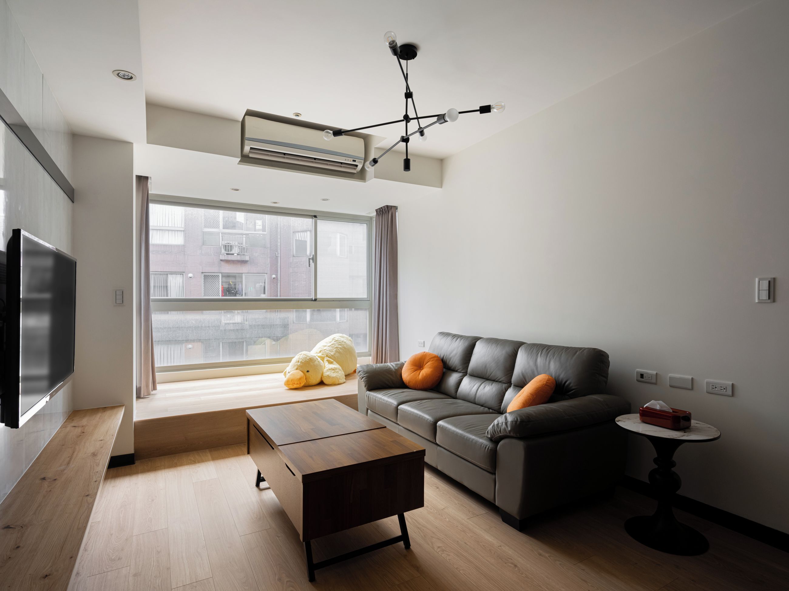 A modern living room with a sofa, a coffee table, a TV, and a large window offering a view of buildings outside.