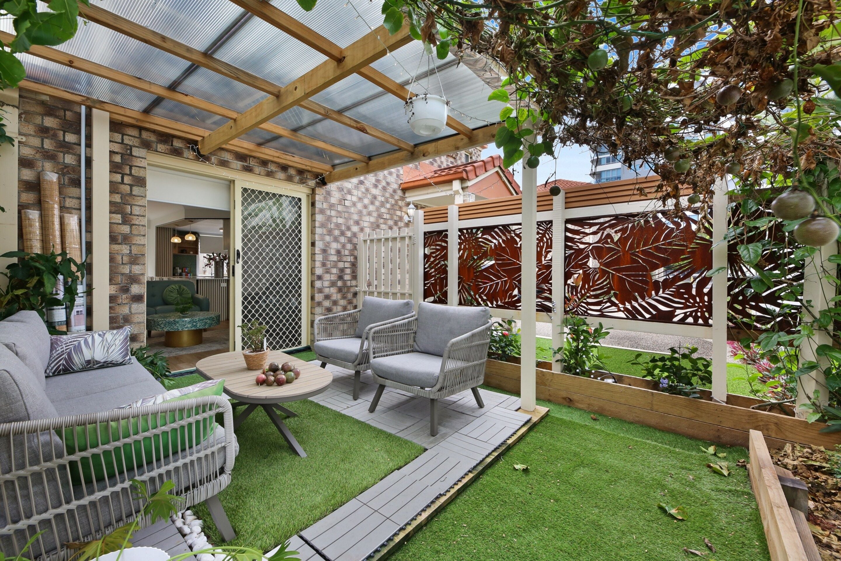 A covered patio area with a seating arrangement, artificial grass, and a view of a brick wall and a building in the background.