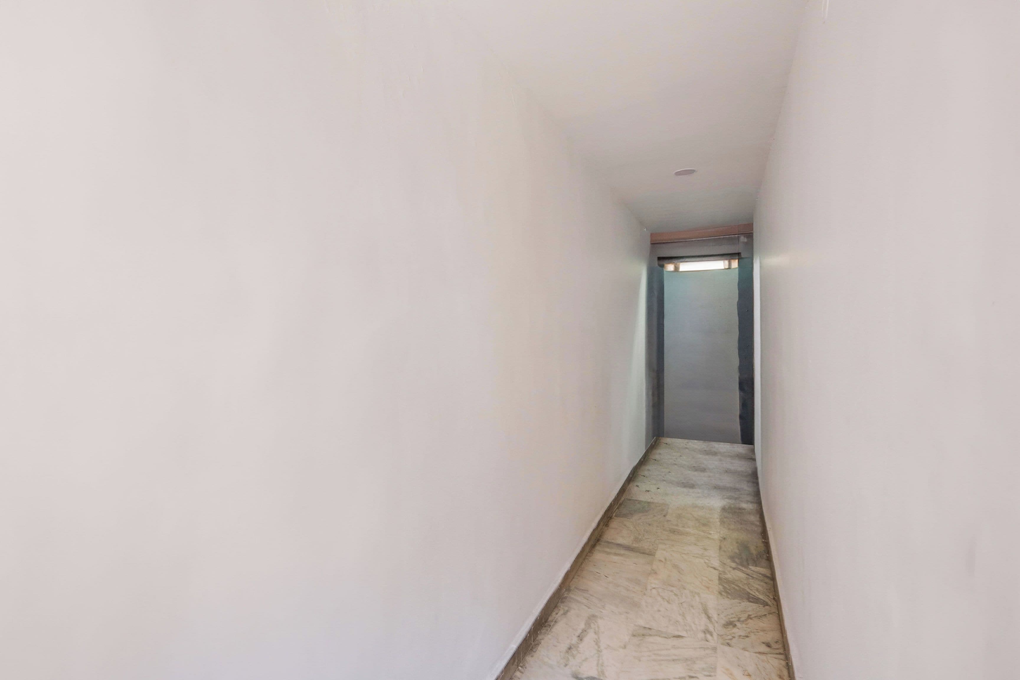 A narrow hallway with white walls and a marble floor leading to a door.