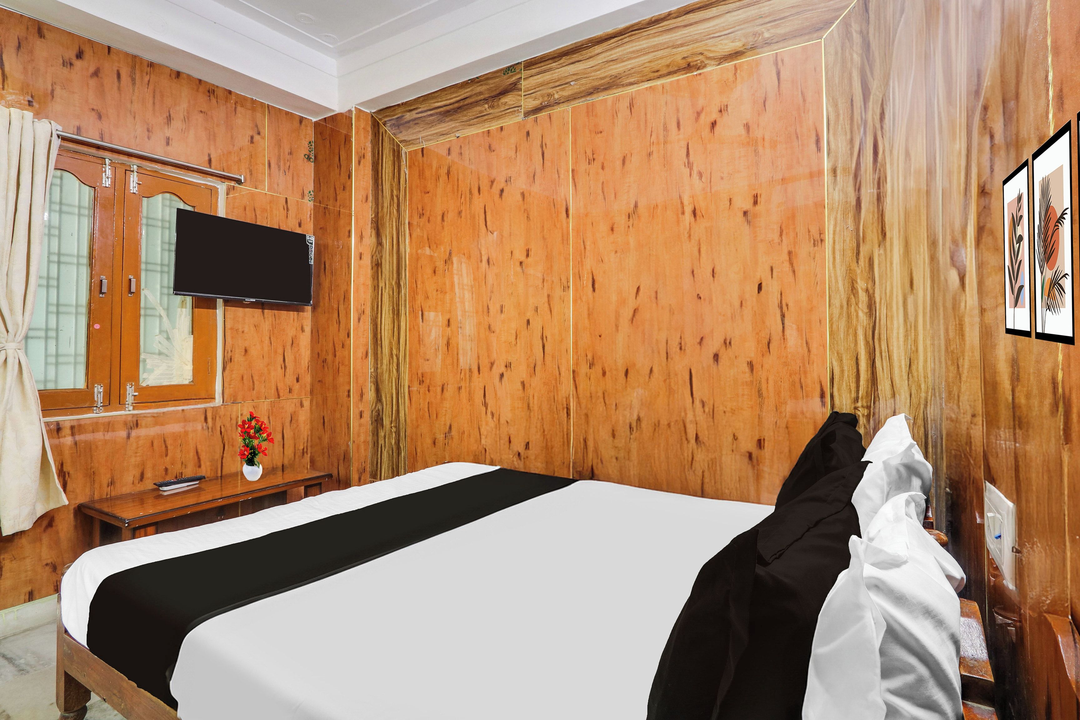 Quarto de hotel com parede de madeira, cama com roupa de cama branca e preta, televisão de tela plana na parede, escrivaninha de madeira com um vaso de flores e uma janela com cortinas.