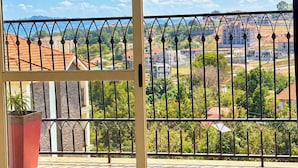 A room with a large window overlooking a cityscape, a balcony with a metal railing, and a bed with a blue quilt. - Serene Harmony Homes (Kampala)