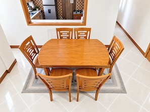 Un coin repas avec une table et des chaises en bois, un tapis et une cuisine visible à travers une fenêtre.