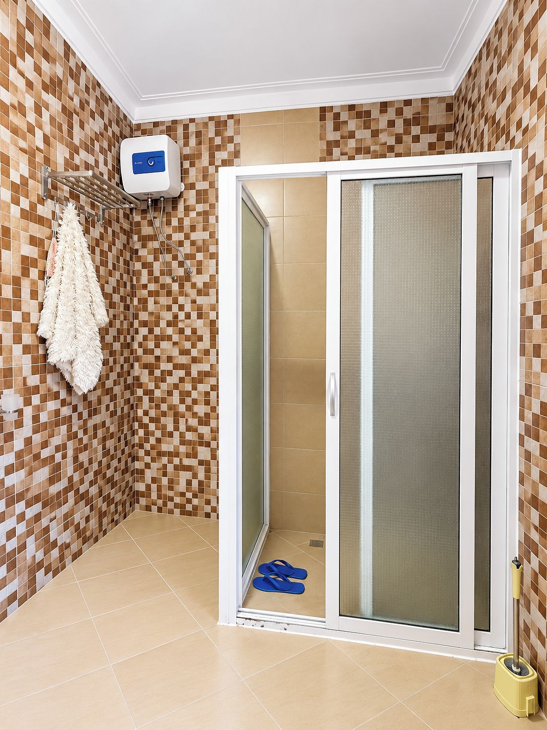 A bathroom with a checkered tile wall, a white towel rack, a shower with a glass door, and a yellow cleaning tool on the floor.