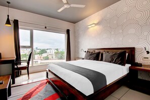 A modern bedroom with a large bed, a desk, and a chair. There is a view of the city through the window. - Super Townhouse Oak Electronic City (Anekal)