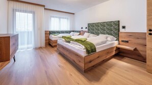Ein modernes Hotelzimmer mit zwei Holzbetten, einer Holzkommode, einem Schreibtisch und einem großen Fenster mit durchscheinenden Vorhängen.