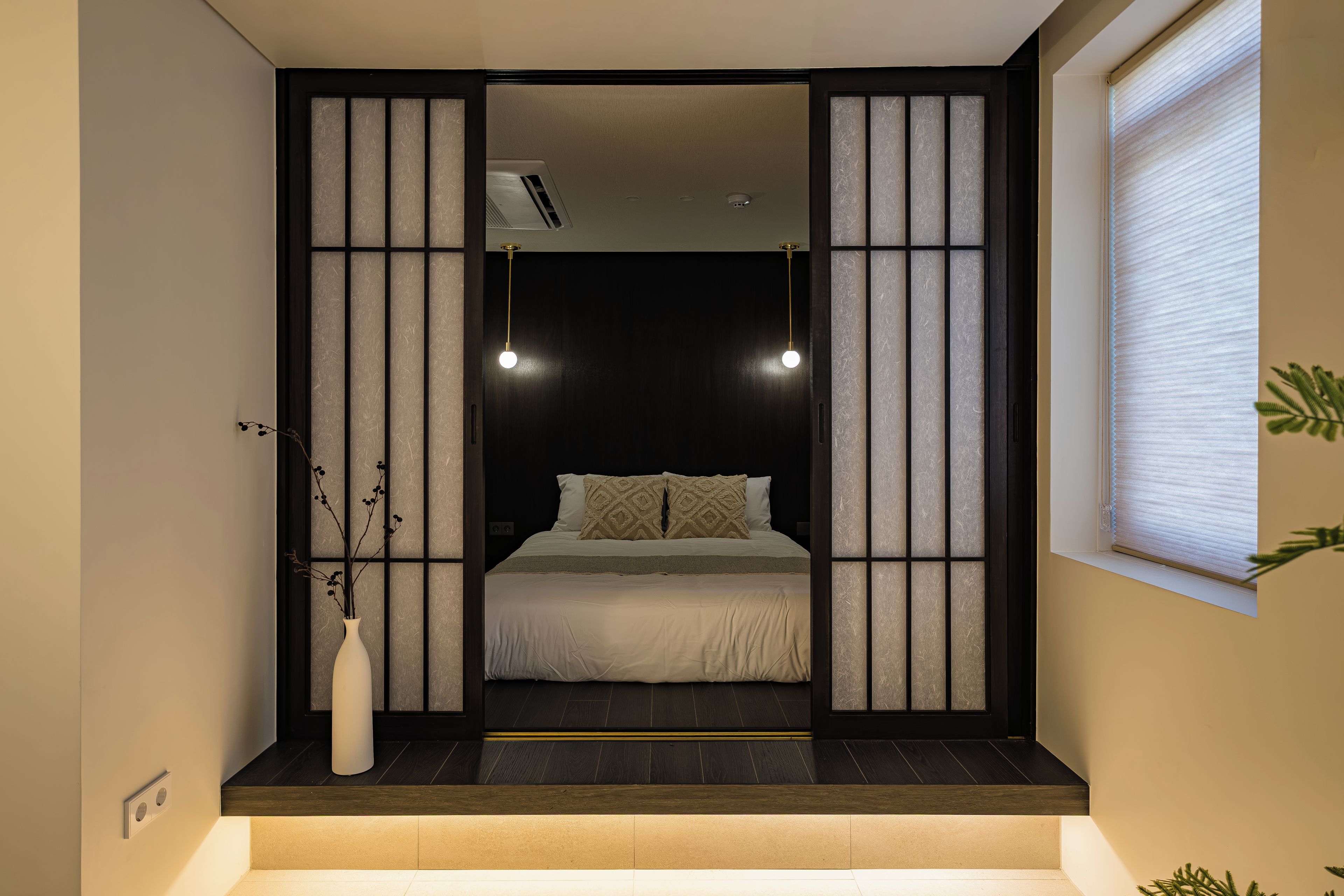 Quarto com cama, mesa de cabeceira, um vaso com galhos e janela com persianas.