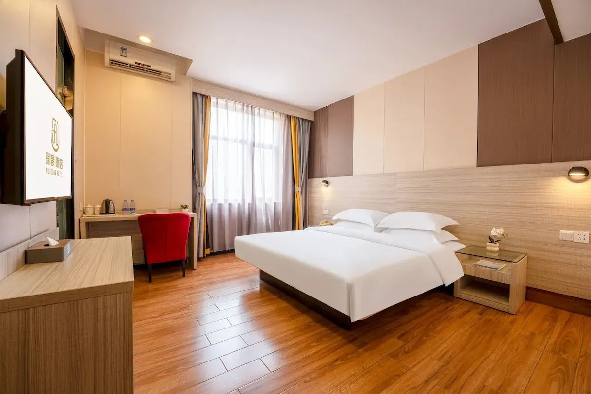 A hotel room with a bed, bedside table, desk, chair, and a large window with curtains.