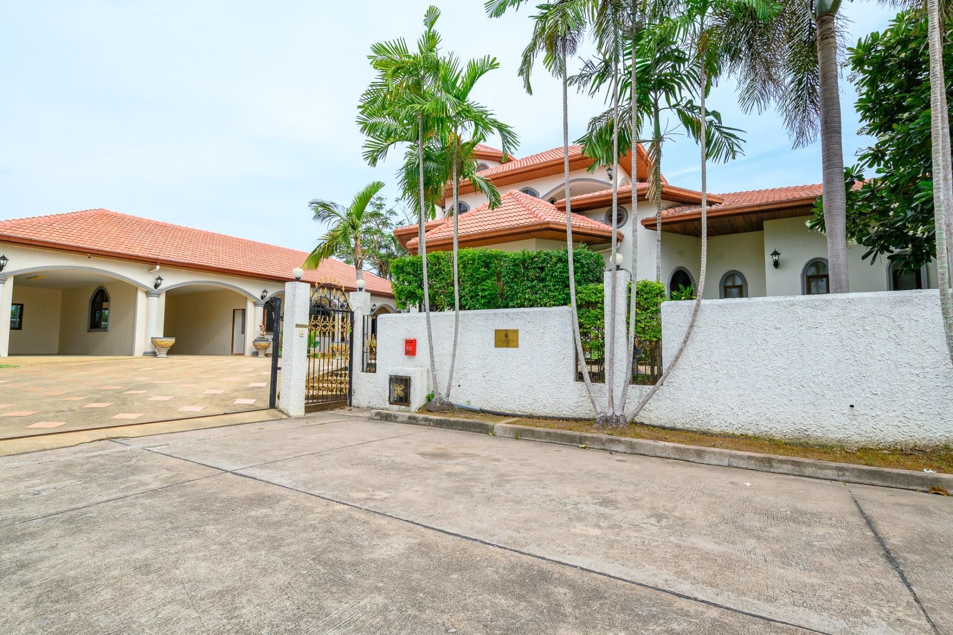Área residencial murada com entrada pavimentada, palmeiras e um edifício com telhado de telhas vermelhas.