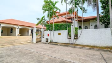 Área residencial murada com entrada pavimentada, palmeiras e um edifício com telhado de telhas vermelhas.