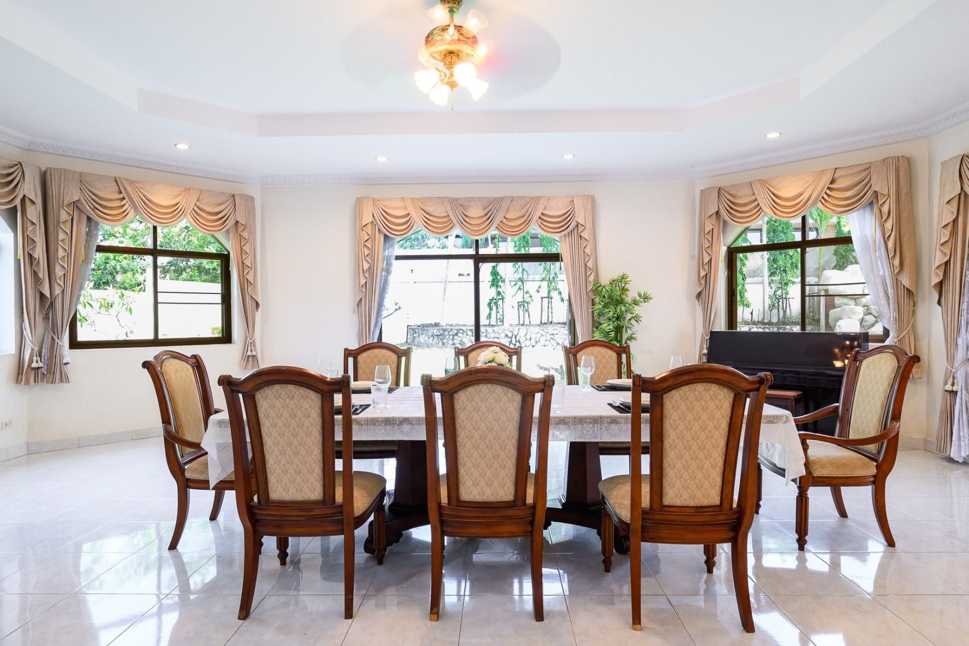 A dining area with a piano, six chairs, and large windows with curtains.