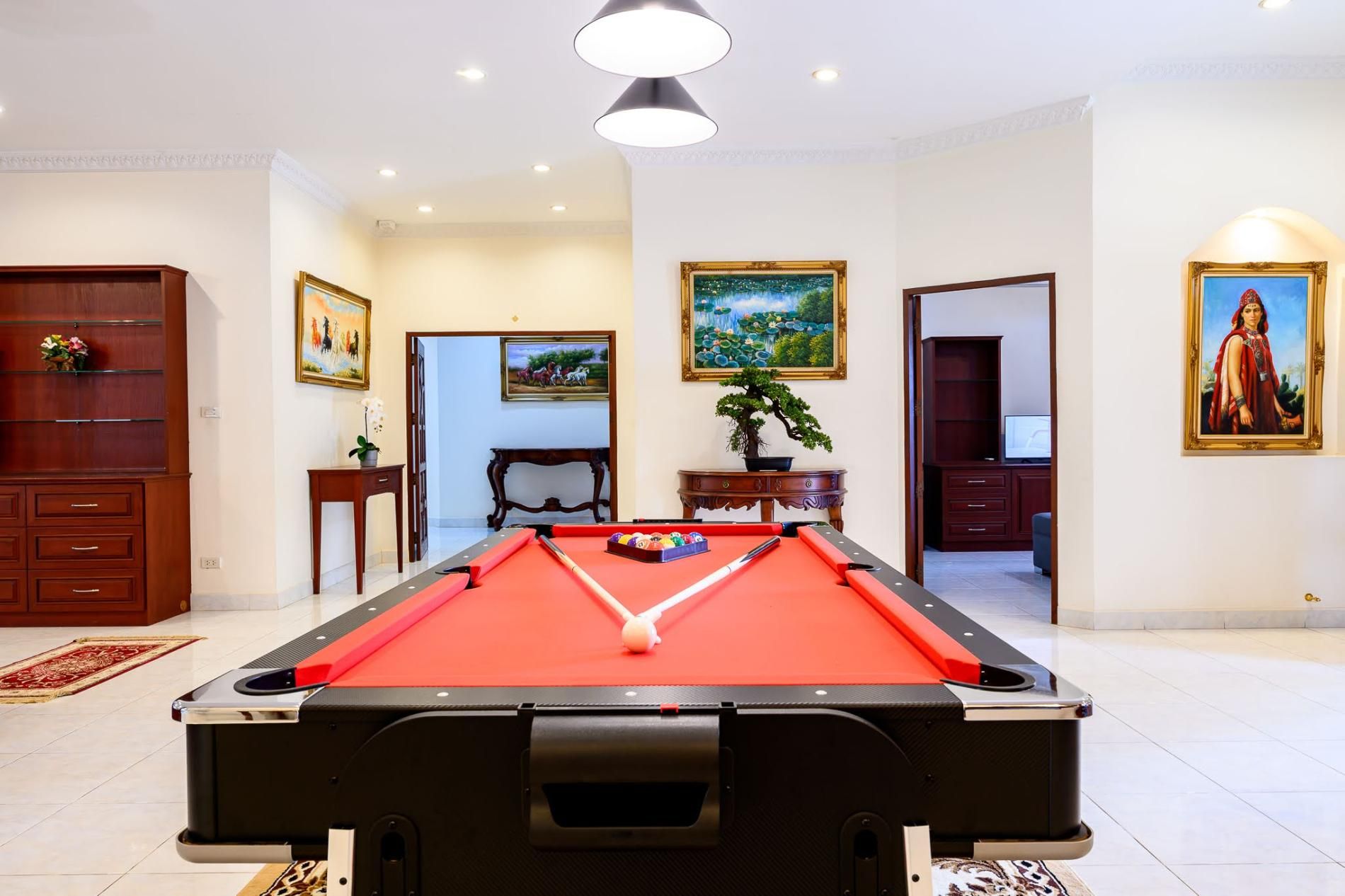 A pool table in a room with a red felt surface, surrounded by walls with framed artwork, a potted plant, and a wooden cabinet.