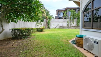 Un patio trasero residencial con césped, un árbol, una pared blanca y una casa con ventana.