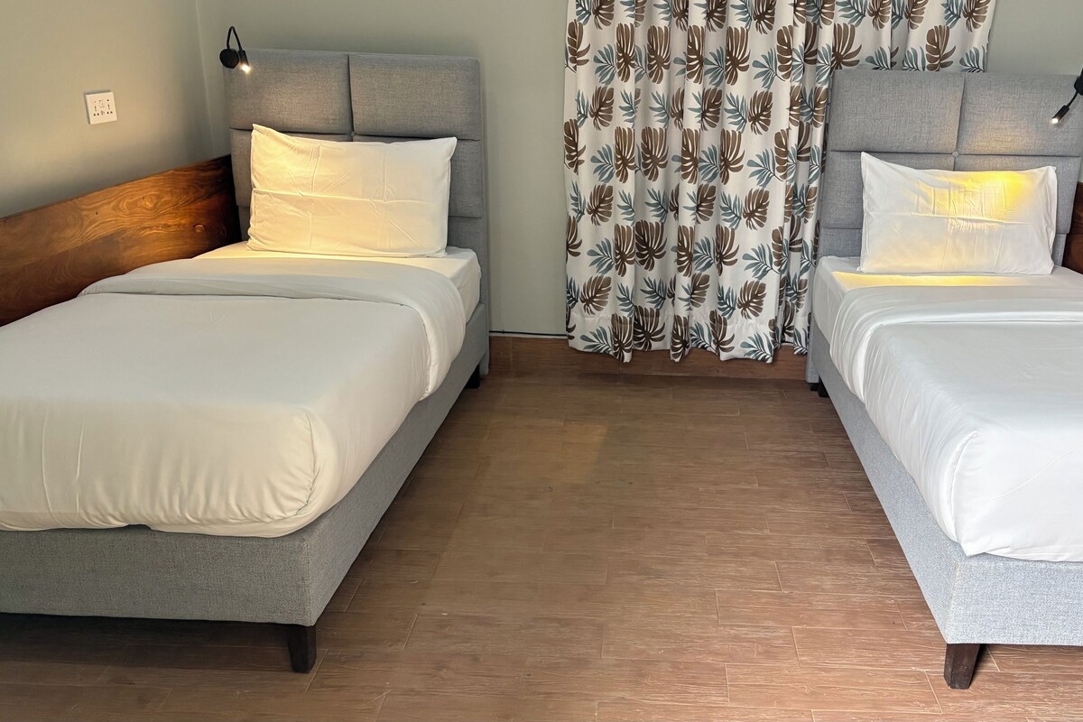 Habitación doble con cabeceros grises, ropa de cama blanca y cortina estampada.