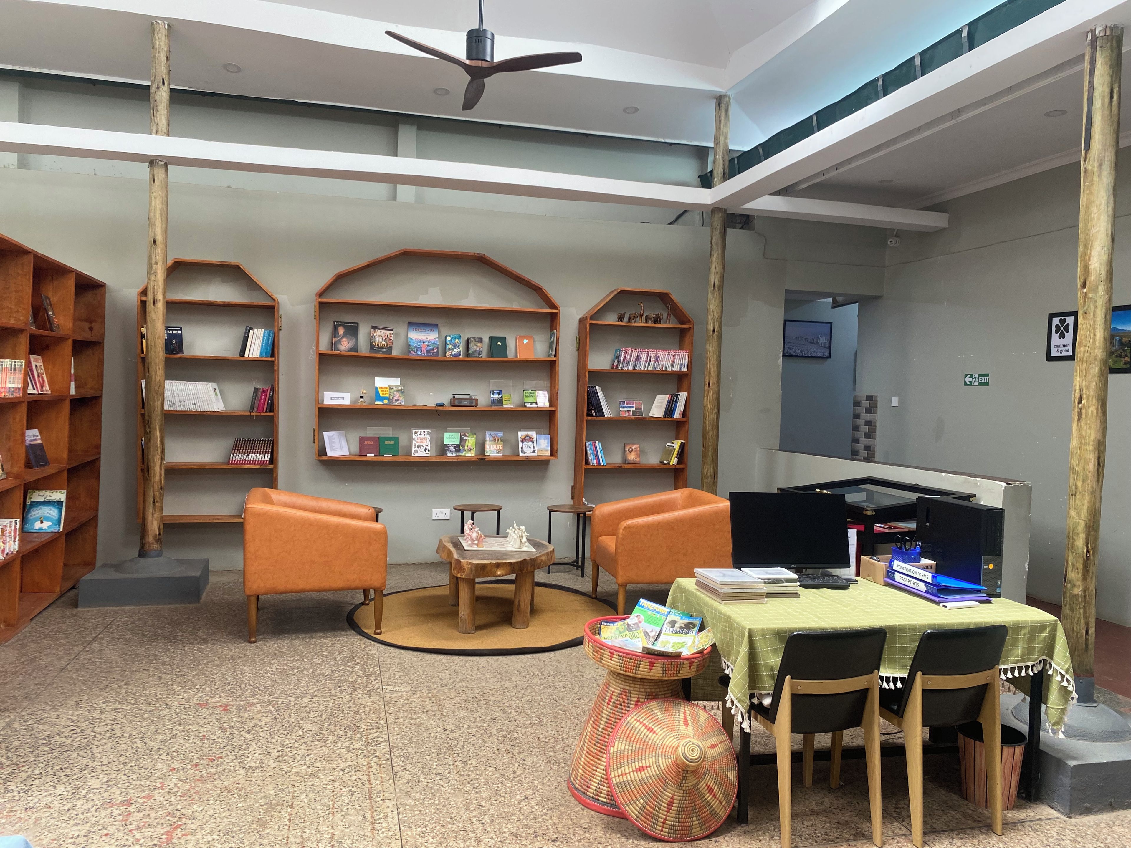 Une bibliothèque avec des étagères, deux fauteuils orange, une table recouverte d’une nappe verte et un ventilateur de plafond.
