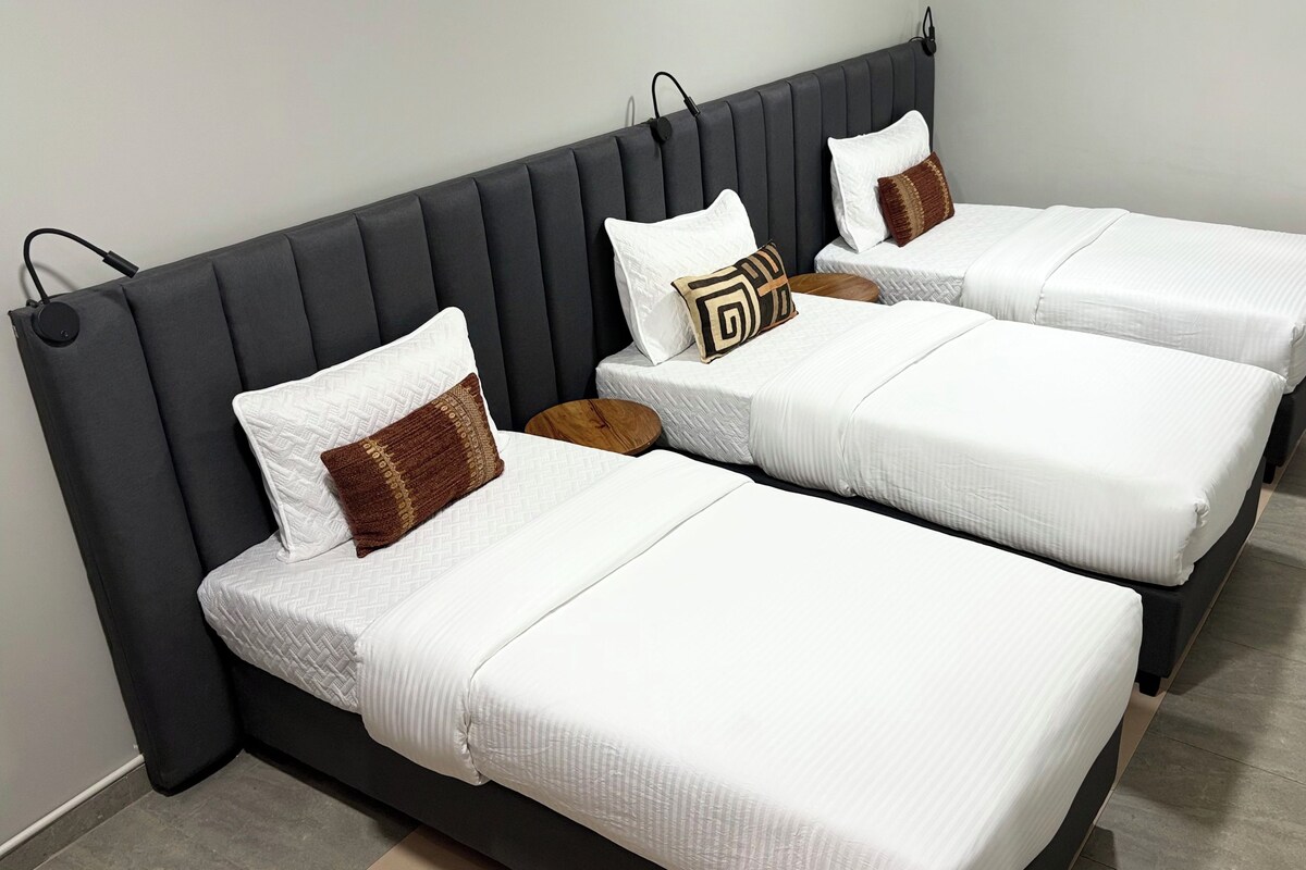 A hotel room with three single beds, each with white bedding and decorative pillows.