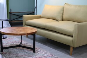 Un sofá color beige con un cojín a juego, una mesa redonda de madera con patas metálicas y una alfombra estampada sobre suelo de baldosas.