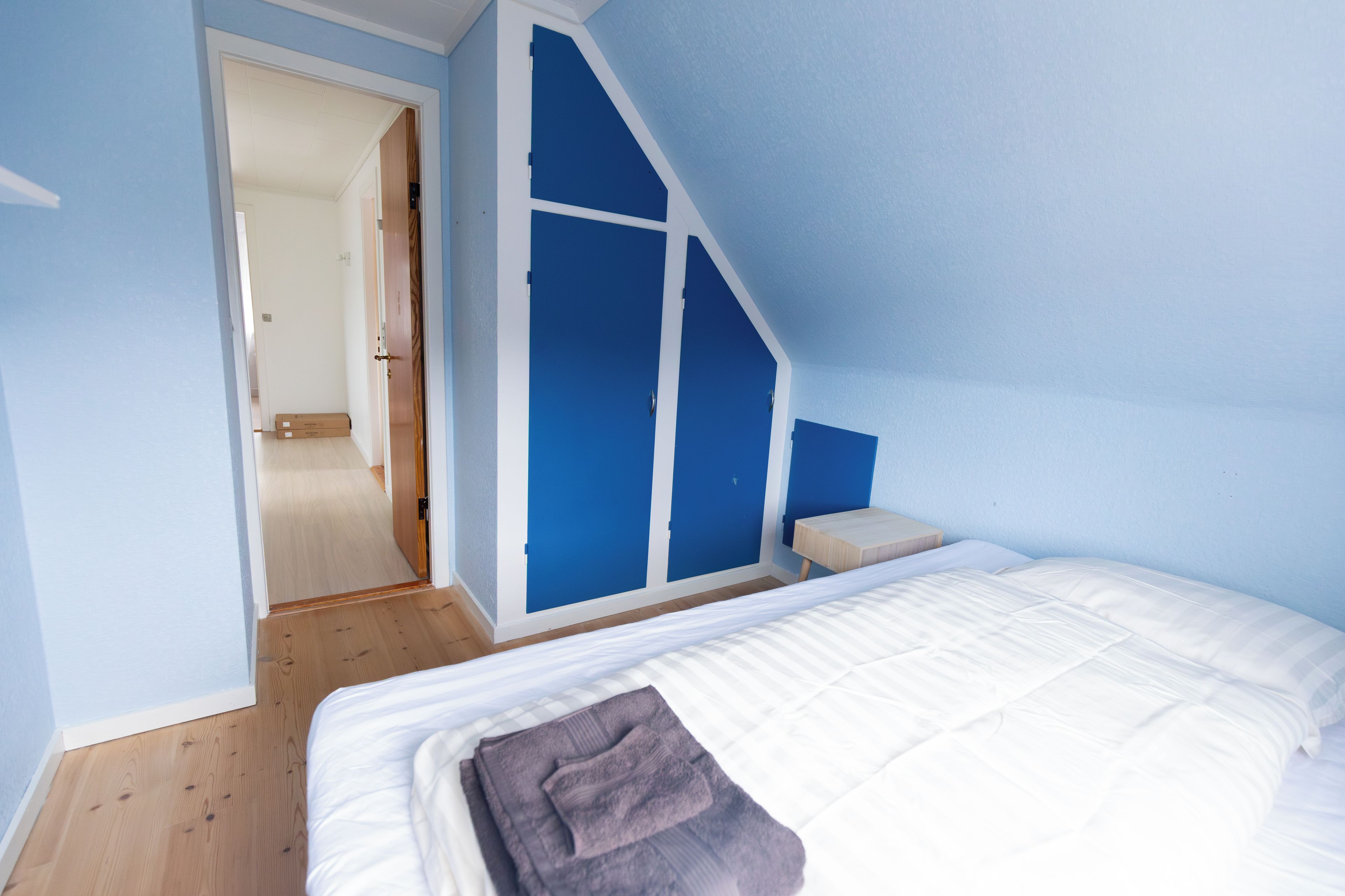 Un dormitorio con una cama, piso de madera, pared azul y una puerta que da a otra habitación.
