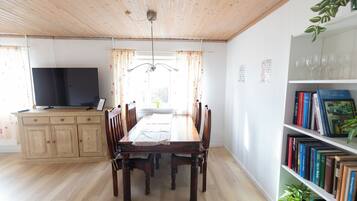 A dining area with a wooden table and chairs, a television on a cabinet, and a bookshelf with books and a plant.