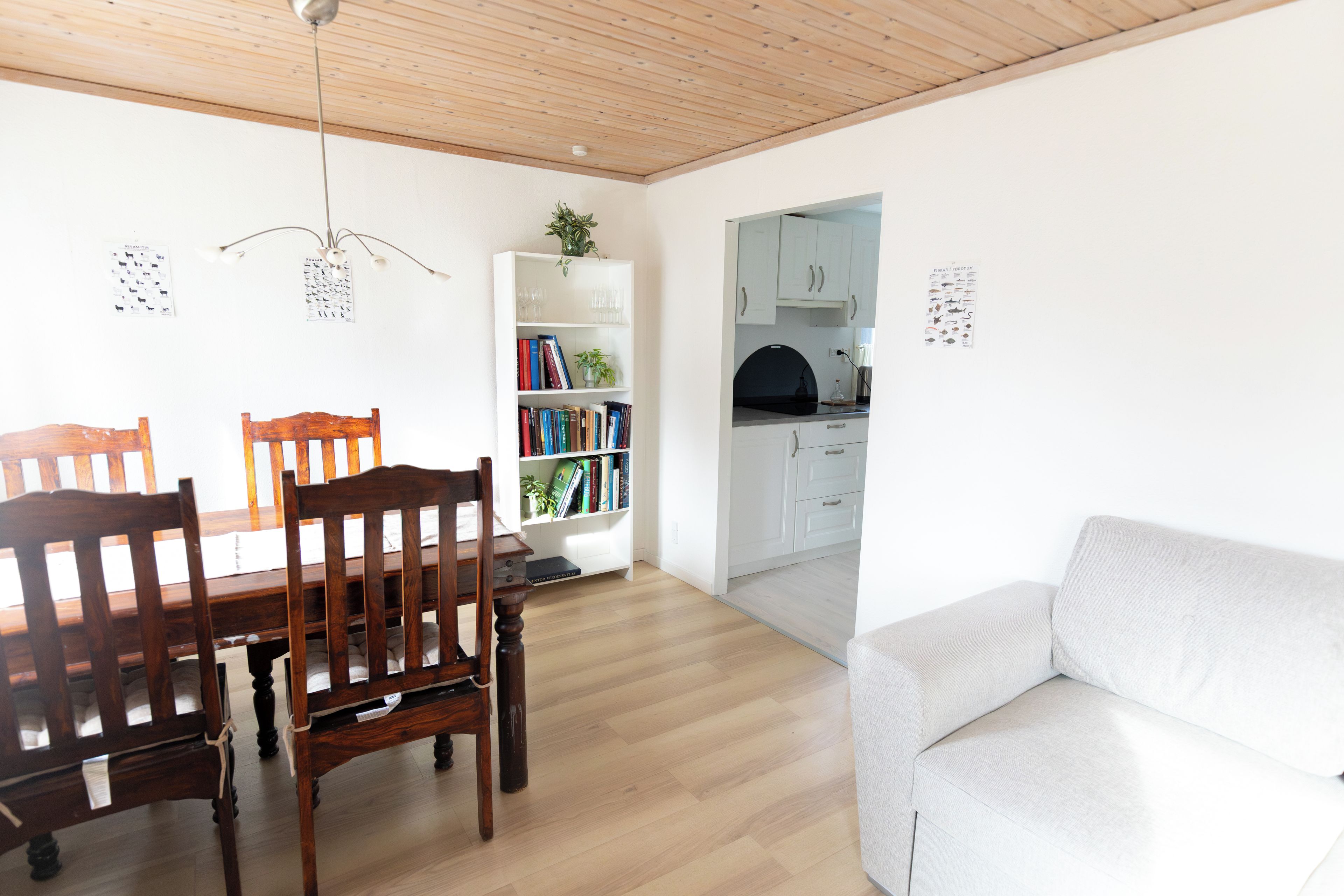 Una sala de estar moderna con comedor, un sofá blanco, sillas de madera, una estantería y una cocina visible a través de un vano abierto.
