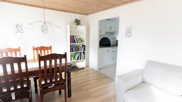 Una sala de estar moderna con comedor, un sofá blanco, sillas de madera, una estantería y una cocina visible a través de un vano abierto.