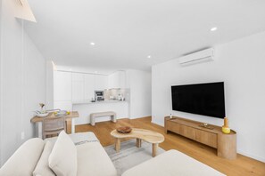 A modern living room with a flat-screen TV, a sofa, a coffee table, and a kitchen area in the background. - Douro Atlântico II | Terrace & Sea Views (Canidelo)