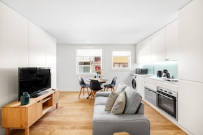 Eine moderne Küche mit weißen Schränken, ein Essbereich mit Tisch und Stühlen sowie ein graues Sofa mit gemusterten Kissen.